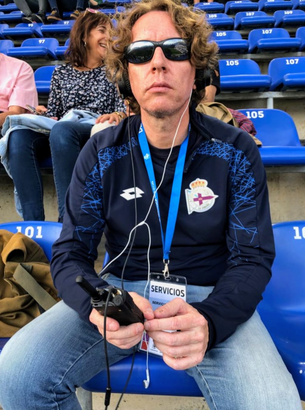 La imagen muestra a Jesús Suárez sentado en las gradas del estadio de Riazor, centrado en primer plano. Con cabello rizado, gafas oscuras y una expresión seria y enfocada, Viste una chaqueta deportiva del Deportivo de La Coruña y lleva un lanyard con una identificación de "SERVICIOS". Con auriculares puestos y sosteniendo un dispositivo electrónico, se le ve involucrado en la comunicación o narración del evento. En el fondo, otras personas se ven en las gradas, desenfocadas, lo que refuerza la atmósfera de un entorno deportivo.