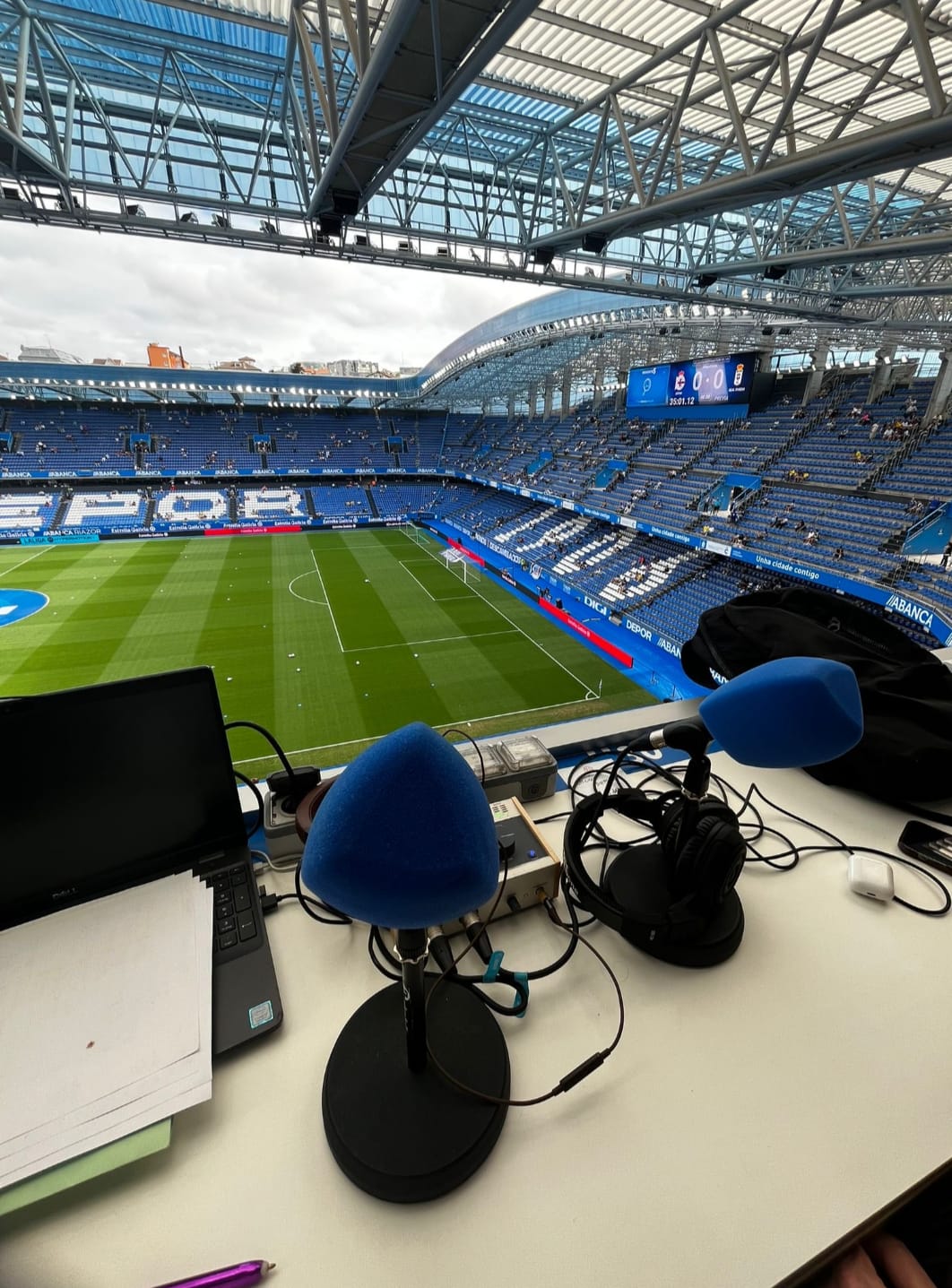 La imagen captura una vista panorámica desde una cabina de retransmisión en el estadio de Riazor, hogar del Deportivo de La Coruña. En primer plano, se observan micrófonos con protectores azules, un ordenador portátil, y varios equipos de transmisión, reflejando la preparación para un evento deportivo. Al fondo, se aprecia el campo de fútbol y las gradas en azul y blanco, parcialmente ocupadas, lo que sugiere que el partido aún no ha comenzado. El moderno techo del estadio y la pantalla gigante añaden una atmósfera profesional y anticipatoria antes del inicio del juego.