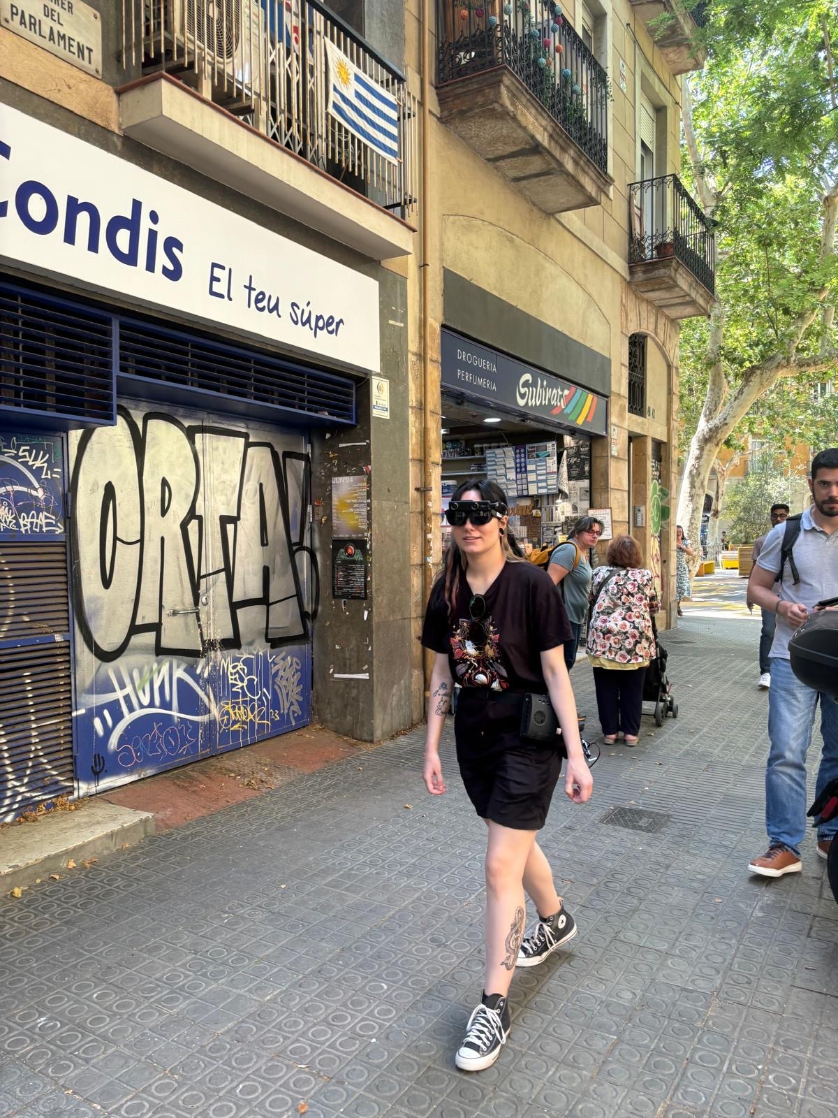 La imagen muestra una calle urbana con varias personas caminando. En primer plano, hay una mujer joven con cabello largo y oscuro, vestida con una camiseta negra, falda corta negra y zapatillas deportivas negras. Lleva unas gafas Biel Smartgaze y tiene tatuajes visibles en las piernas. A su alrededor, se pueden ver otras personas caminando, algunas con carritos de bebé. A la izquierda, hay un edificio con un letrero que dice "Condis El teu súper" y una persiana metálica con grafitis. En el balcón del edificio, hay una bandera de Uruguay. A la derecha, hay una tienda con el letrero "Droguería Perfumería Subirà". Al fondo, se ven árboles que proporcionan sombra a la calle.